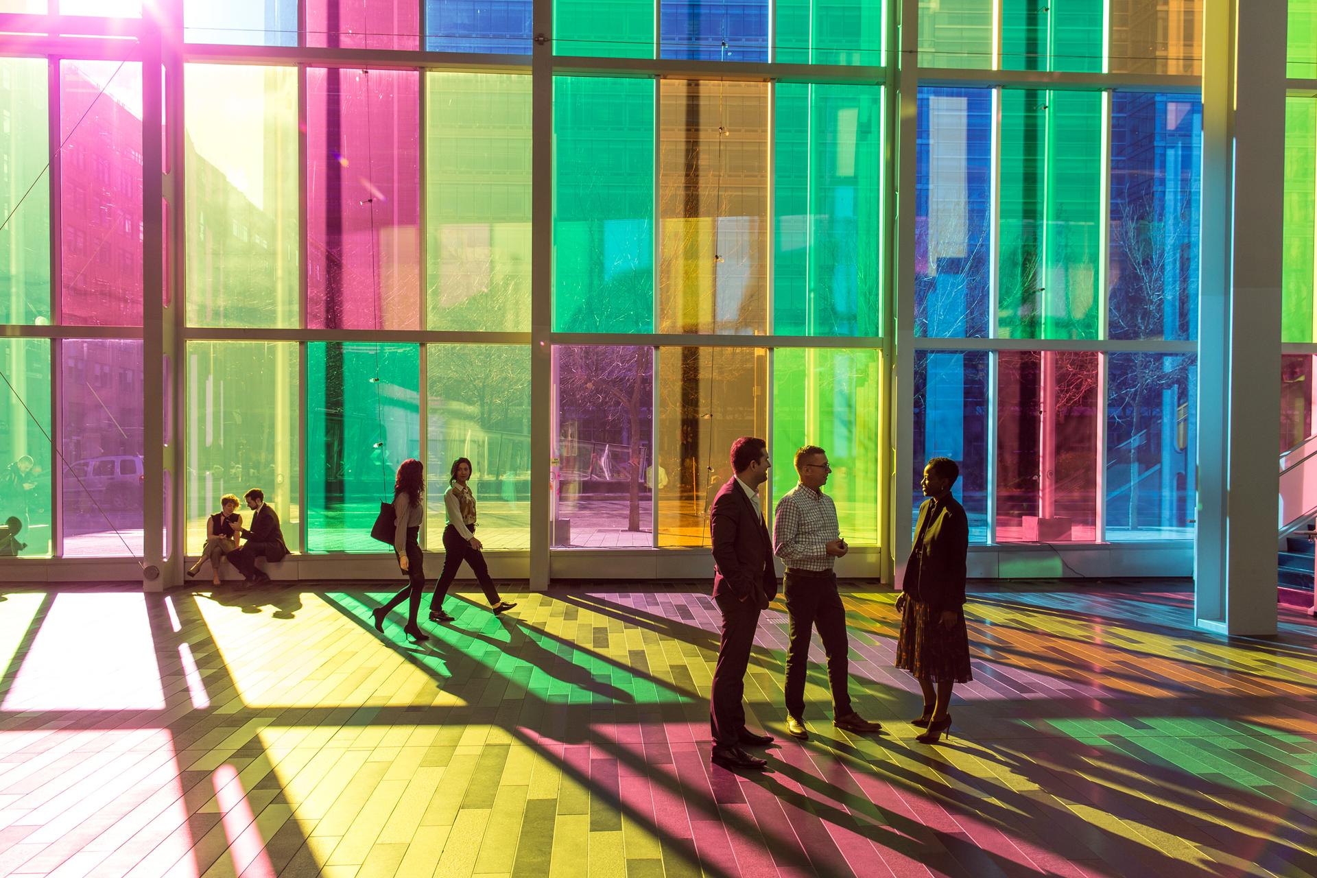 Des groupes de gens se trouvent dans un large couloir où la lumière filtre à travers des panneaux de verre coloré.