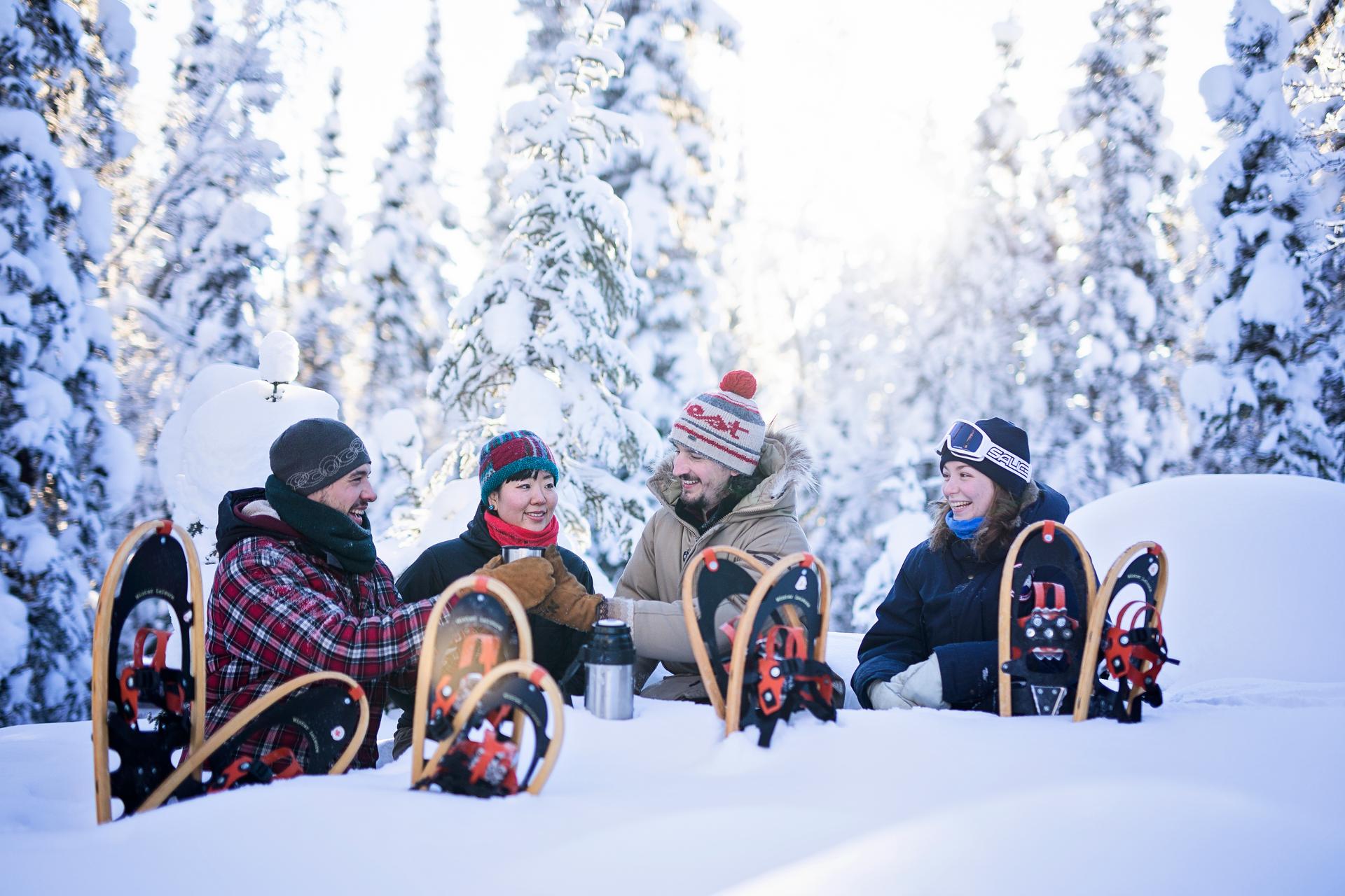 Four people snowshoeing in a snowy forest, sharing a warm drink surrounded by snow-covered trees